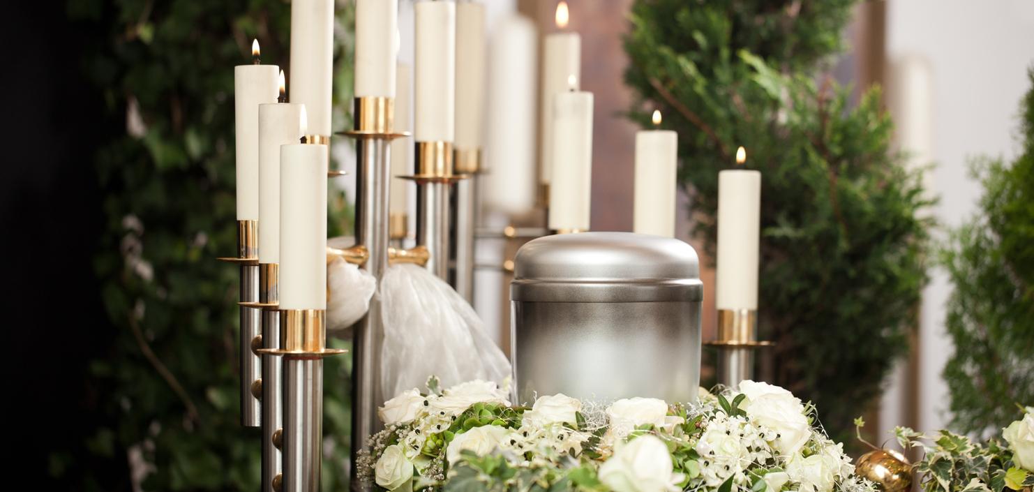 Silberne Urne umgeben von weißen Rosen und Kerzen auf einem Tisch, im Hintergrund grüne Pflanzen.