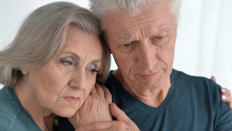 Ein älteres Paar steht eng umschlungen, beide blicken nachdenklich und wirken emotional berührt.