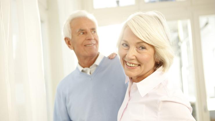 Ein lächelndes älteres Paar steht vor einem Fenster, umgeben von hellem Licht, mit sanften Farben im Hintergrund.