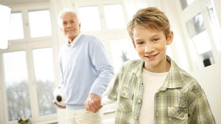 Ein Junge lächelt, während er die Hand eines älteren Mannes hält, in einem hellen Raum mit Fenstern.